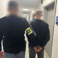 Policjant prowadzi zatrzymanego mężczyznę korytarzem komendy; twarze są celowo zamazane.