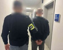 Policjant prowadzi zatrzymanego mężczyznę korytarzem komendy; twarze są celowo zamazane.