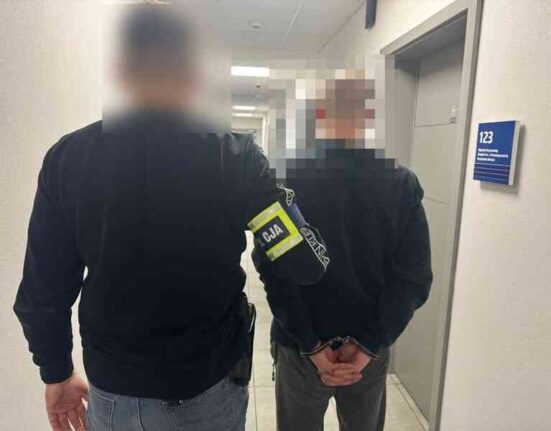 Policjant prowadzi zatrzymanego mężczyznę korytarzem komendy; twarze są celowo zamazane.