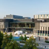 Widok z zewnątrz na Galerię Młociny w Warszawie z budynkiem centrum handlowego i strefą rekreacyjną