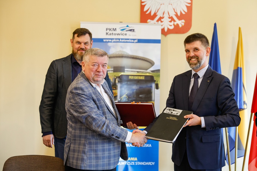 Podpisanie umowy między PKM Katowice a firmą Solaris Bus & Coach na dostawę 15 autobusów CNG. Dwóch mężczyzn ściska dłonie trzymając teczkę z dokumentami, w tle rollup PKM Katowice i flagi.